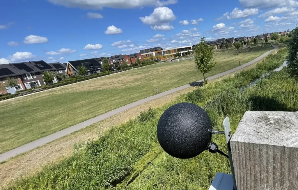 Grasveld met huizen, waarbij een zwarte bolsensor hangt in een paal om de WGBT te meten.